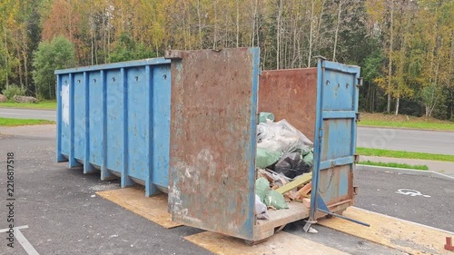 Open blue dumpster filled with garbage bags and construction debris. Rusty metal container on a paved lot, autumn trees, overcast day.