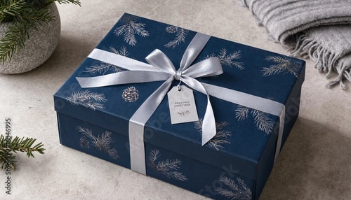Navy blue Christmas gift box with silver bow and gift tag. Pine branch print present on a light stone surface with fir twigs and a pine cone, ideal for luxury winter holiday and packaging projects.