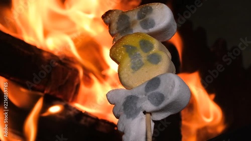 Halloween ghosts and pumpkin-shaped marshmallows roasting and melting on a wooden skewer over bright orange campfire flames during a festive autumn night outdoors