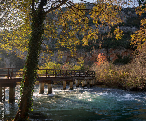 Wooden footbridge over rushing turquoise river water surrounded by autumn trees and limestone cliff, Krka National Park, Croatia