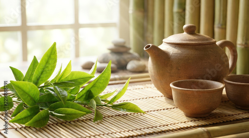 Fresh camellia sinensis tea leaves with clay teapot and cups on bamboo mat in bright natural sunlight for wellness and zen lifestyle