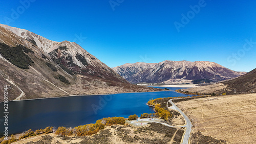 Aerial view of Lake Pearson in Arthur's Pass, featuring blue water, rugged mountains, golden autumn trees, and a winding road, Arthur's Pass, Canterbury Region, New Zealand.