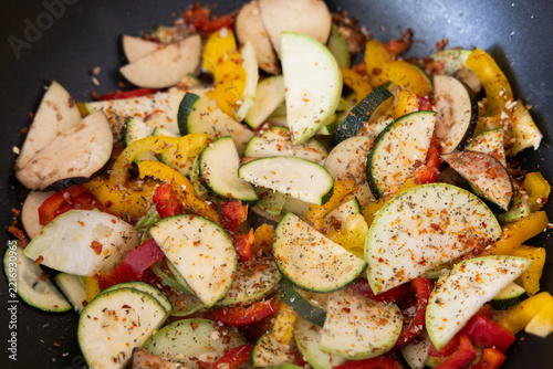 Sliced zucchini and bell peppers cooking in a pan with spices against a dark metal background
