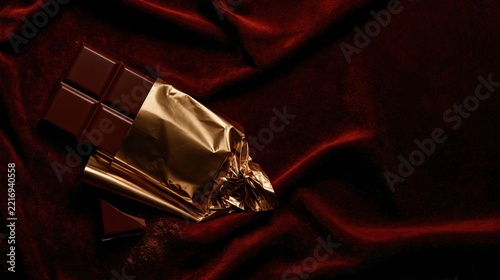 Dark chocolate bar in crinkled gold foil on deep burgundy velvet, a glossy luxury treat with a snapped piece for holiday campaigns and copy space