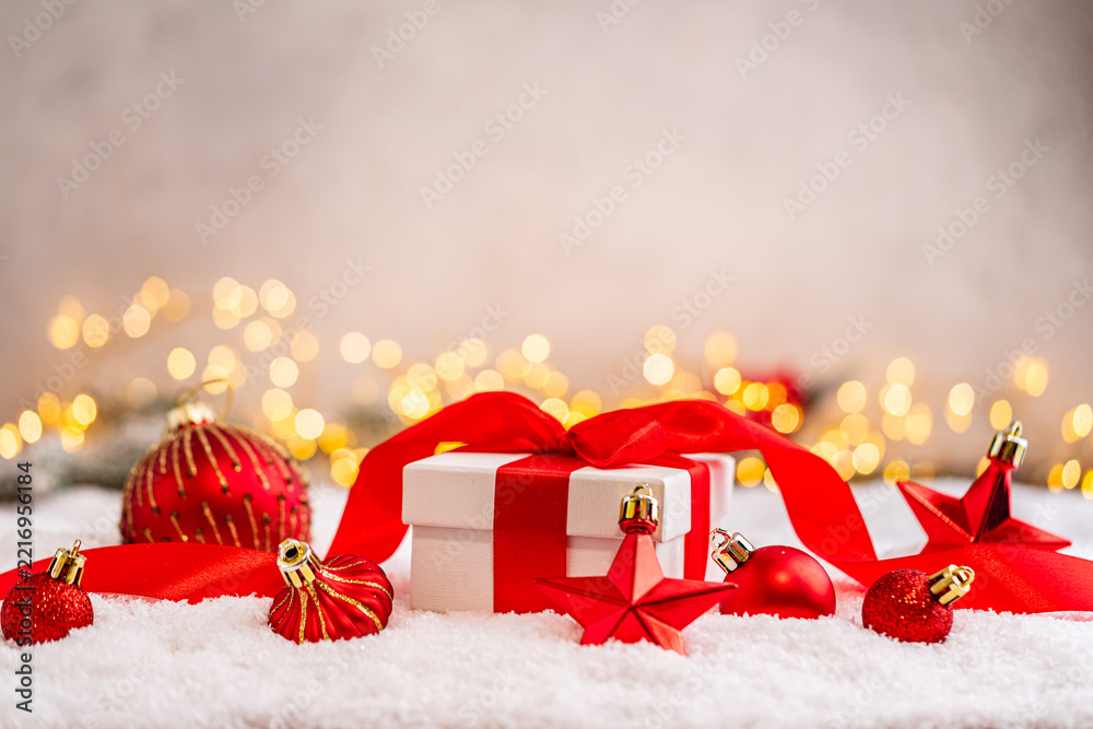 Fototapeta premium A Christmas background featuring a white gift box tied with a red satin bow surrounded by red star and bauble ornaments on snow, softly lit by warm twinkling lights with copy space.