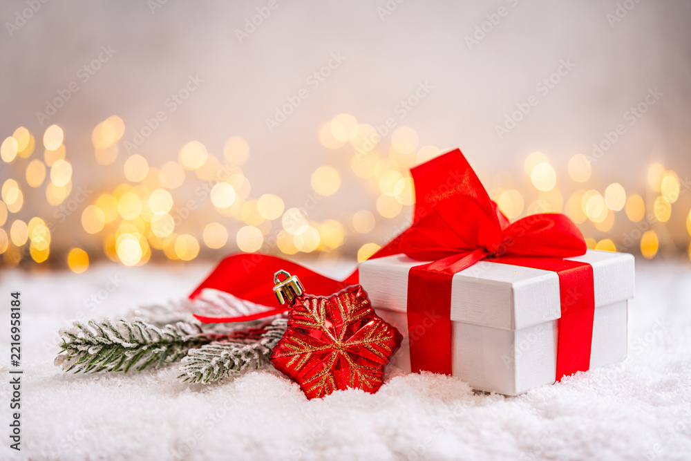 Fototapeta premium A Christmas background featuring a white gift box tied with a large red satin bow beside a frosted pine branch and red ornament on snow, softly lit by warm twinkling lights with copy space.