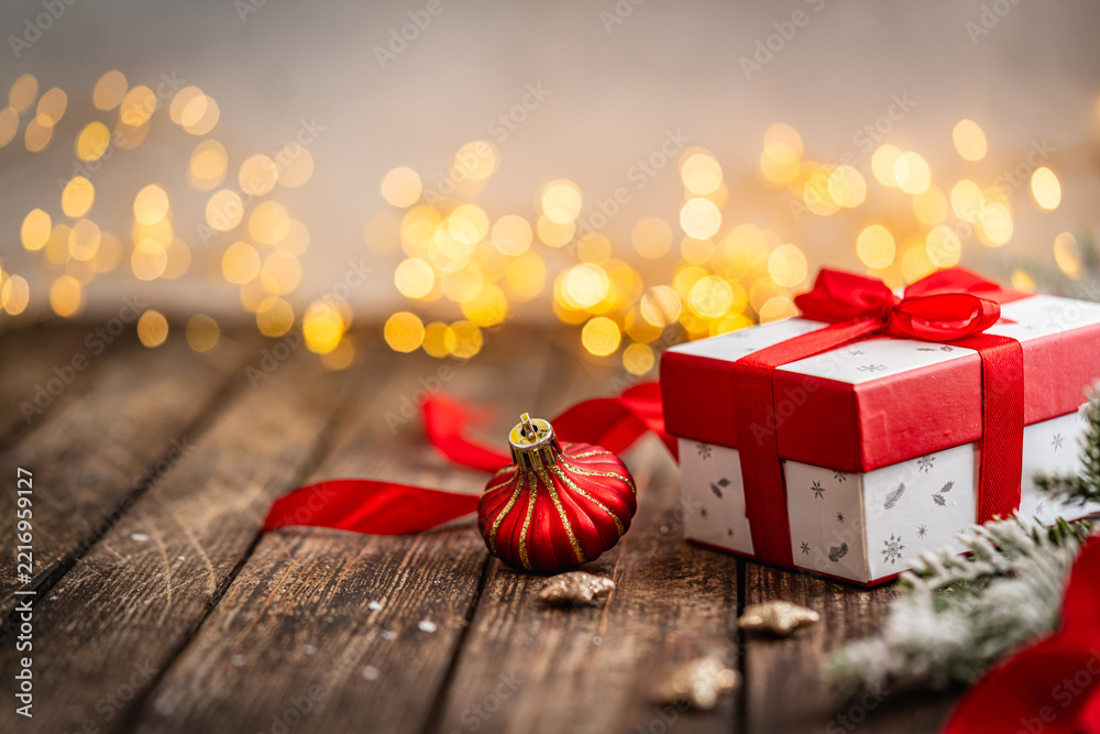 Fototapeta premium A Christmas background featuring a classic white gift box tied with a bright red satin bow beside a golden ornament and frosted pine on rustic wood, warmly lit with twinkling lights.