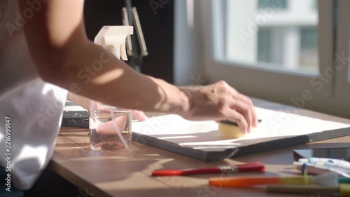 Woman sprays liquid onto painted panel and wipes surface with sponge on worktable