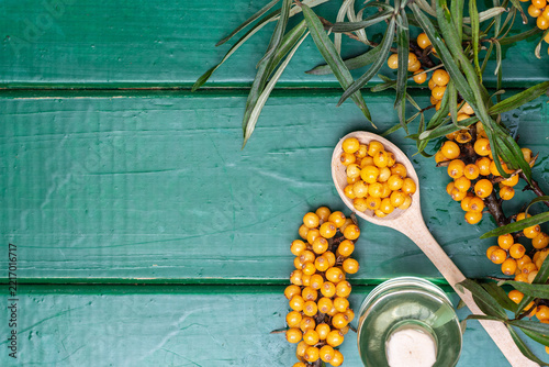  Sea buckthorn berries on branch with oil bottle and wooden spoon on green background. Organic herbal medicine concept for immunity boost