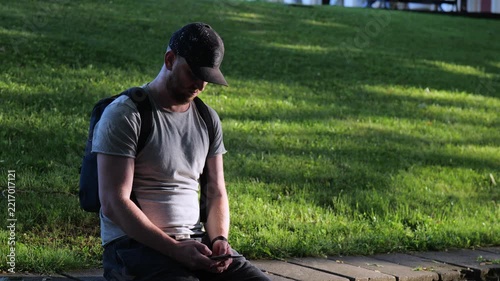 man with backpack using smartphone outdoors in city park video screenshot, side view of young man in cap texting on mobile phone in park, man holding smartphone and typing message outdoors video