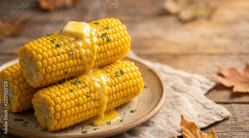 Cooked yellow maize cobs covered in glossy melted dairy spread and chopped parsley rest on earthy platter, surrounded by rustic table settings that evoke inviting harvest dinner atmosphere.