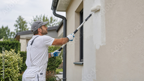 Painter applying fresh paint to an exterior house wall with an extension roller. Exterior painting and home maintenance. Renovation, repair and property upkeep. Painting contractor.