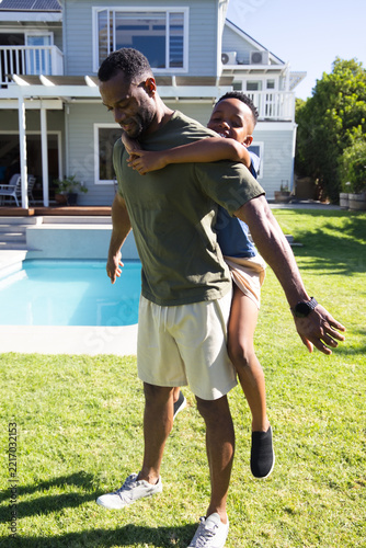 African American father and school-age son playing piggyback on lawn by pool with smartwatch