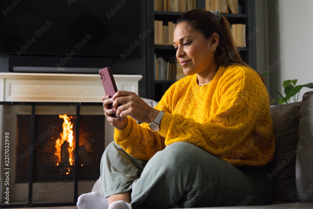 Fototapeta premium Mature African American woman sitting on sofa in living room by fireplace holding pink smartphone