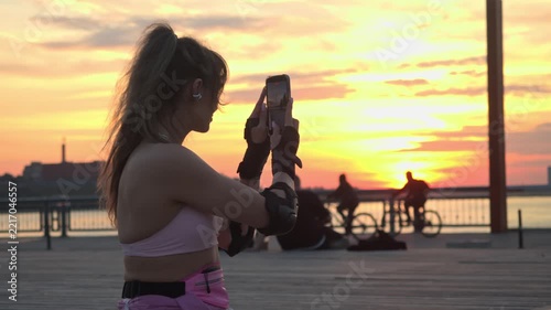 Girl records video using smartphone sitting on wooden promenade. She captures beautiful summer sunset while cyclists ride bikes in background during warm golden hour.