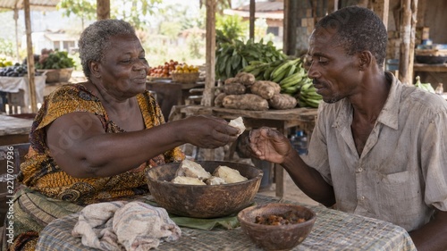 An older woman in a patterned top offers a piece of food to a seated man across a table setting; they are surrounded by market goods like root vegetables and produce