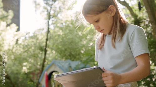 Focused young girl uses digital tablet during backyard leisure play outdoors. Attentive child interacts with touchscreen device while sitting outside under bright natural summer sunlight.