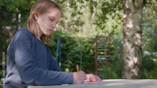 Young girl drawing with pencil on paper sits outdoors at backyard table in bright daylight. Focused student pauses sketching while daydreaming in serene garden during calm summer afternoon.