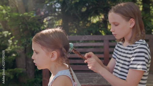 Preteen girl plaits blonde hair of little sister with focused care in lush garden. Caring siblings enjoy relaxed childhood backyard play sitting together near wooden bench in sunny daylight.