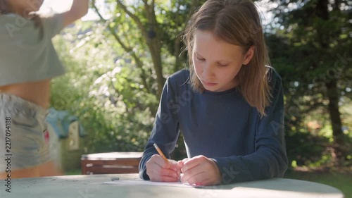 Engaged preteen girl sits outside enjoying homeschooling creative activity on sunny day. Dedicated pupil carefully draws on paper with pencil while younger sister plays behind her in yard.