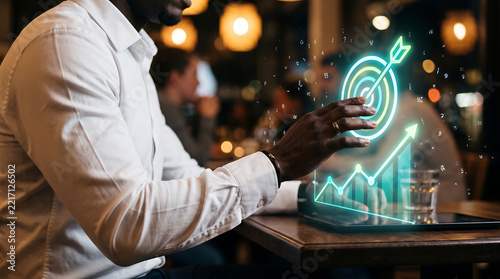 Side view of a man in a white shirt interacting with a glowing neon target and growth chart hologram on a tablet screen in a dimly lit restaurant