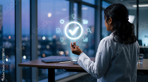Woman in a white lab coat standing in a dark office at night, interacting with a glowing holographic checkmark icon surrounded by digital business symbols