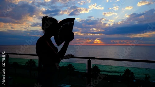 Backlit silhouette of a young woman fanning herself while standing on a balcony, enjoying the beautiful and dramatic sunrise over the calm sea during a relaxing summer