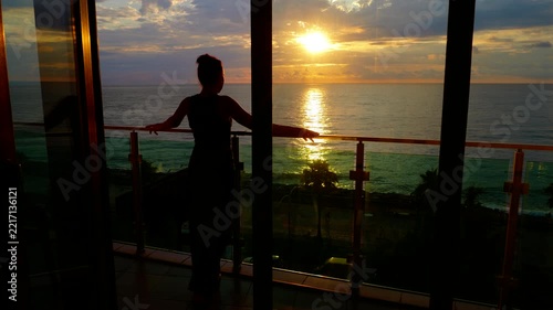 Silhouette of a young woman standing on the balcony of a luxury hotel room, contemplating the amazing golden sunset over the tranquil sea, creating a romantic atmosphere