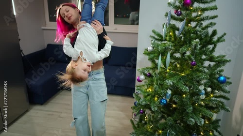 Mother is taking her daughter on hands and turning her upside down standing in living room next to decorated Christmas tree. They are having fun and laughing. Spending time with family concept