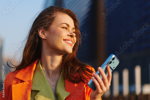 Crypto trader, joyful young woman in profile with open happy smile holds smartphone in sunny city street. Concept of successful mobile trading and digital investment lifestyle.
