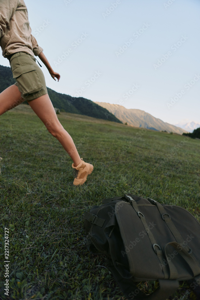 Naklejka premium Young woman in khaki shorts leaps across a sunlit meadow with a backpack on the ground, mountains on the horizon, conveying carefree outdoor adventure and movement.
