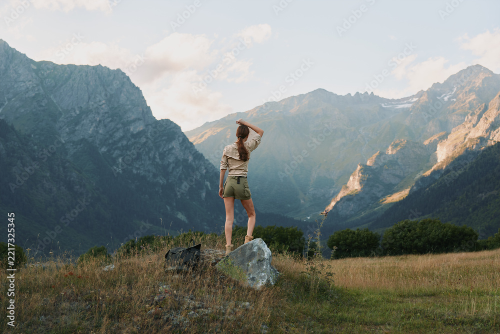 Naklejka premium Young woman stands on a rock in a sunlit mountain meadow, gazing toward distant peaks with a sense of freedom and calm, capturing a natural outdoor moment.