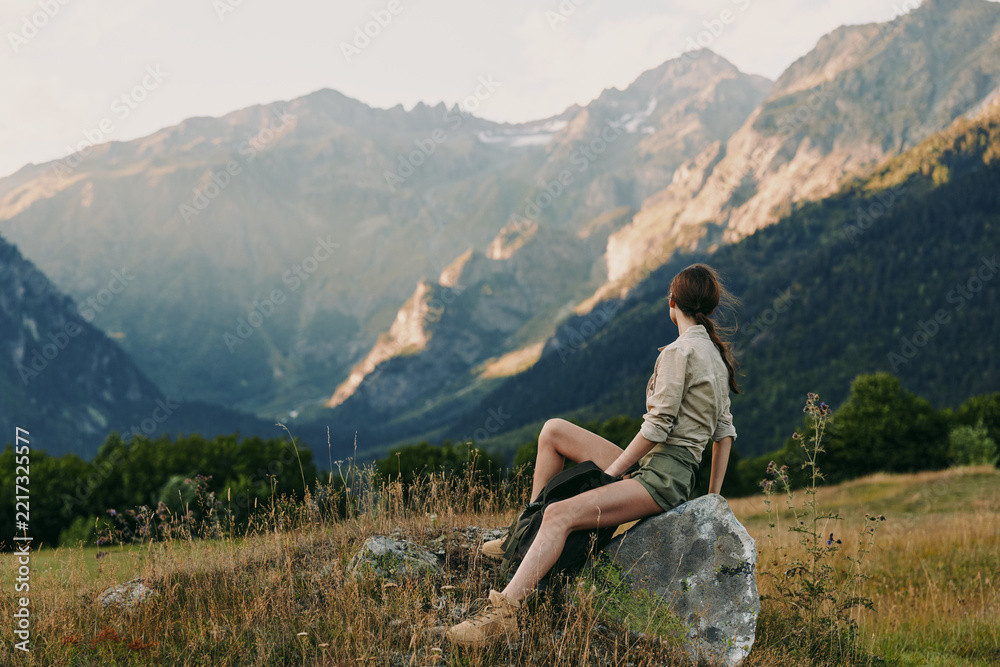 Fototapeta premium One woman sits on a rock in a mountain meadow, gazing toward distant peaks, conveying calm, freedom and solitude in a pristine outdoor scene.