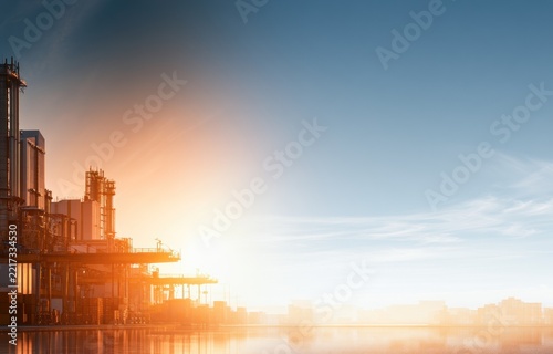 Industrial complex structures reflect on calm water at sunrise with dramatic sky