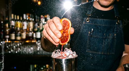 Professional bartender squeezing a fresh blood orange slice into a cocktail shaker with ice.