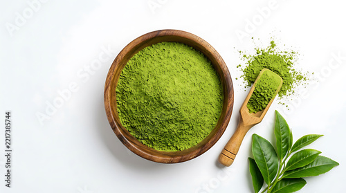 Top view of green matcha tea powder in wooden bowl with scoop, spilled powder, and fresh tea leaves isolated on white background with copy space