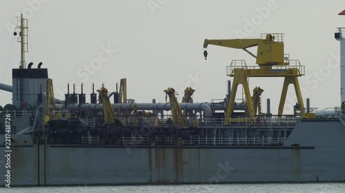 Large dredger ship with yellow deck crane, pipes and white bridge with orange roof anchored on calm river, rusty steel hull and industrial marine equipment under hazy sky