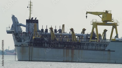 Large dredger ship with yellow deck crane, pipes and white bridge with orange roof anchored on calm river, rusty steel hull and industrial marine equipment under hazy sky