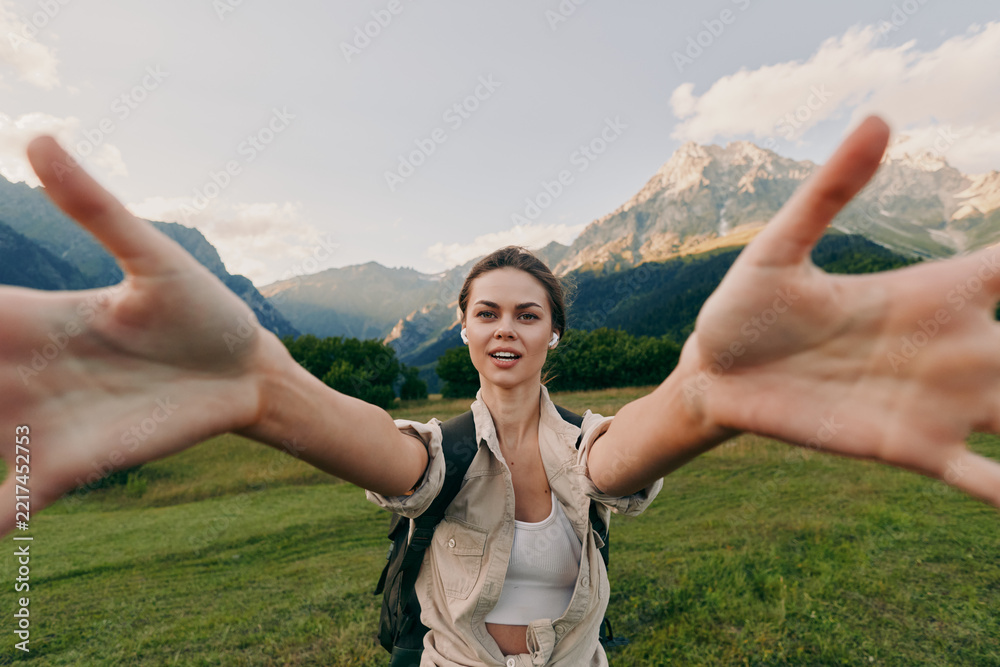 Naklejka premium A young woman in casual clothes stretches her arms forward smiling in a green mountain meadow under a blue sky. Concept for lifestyle, nature, and freedom.