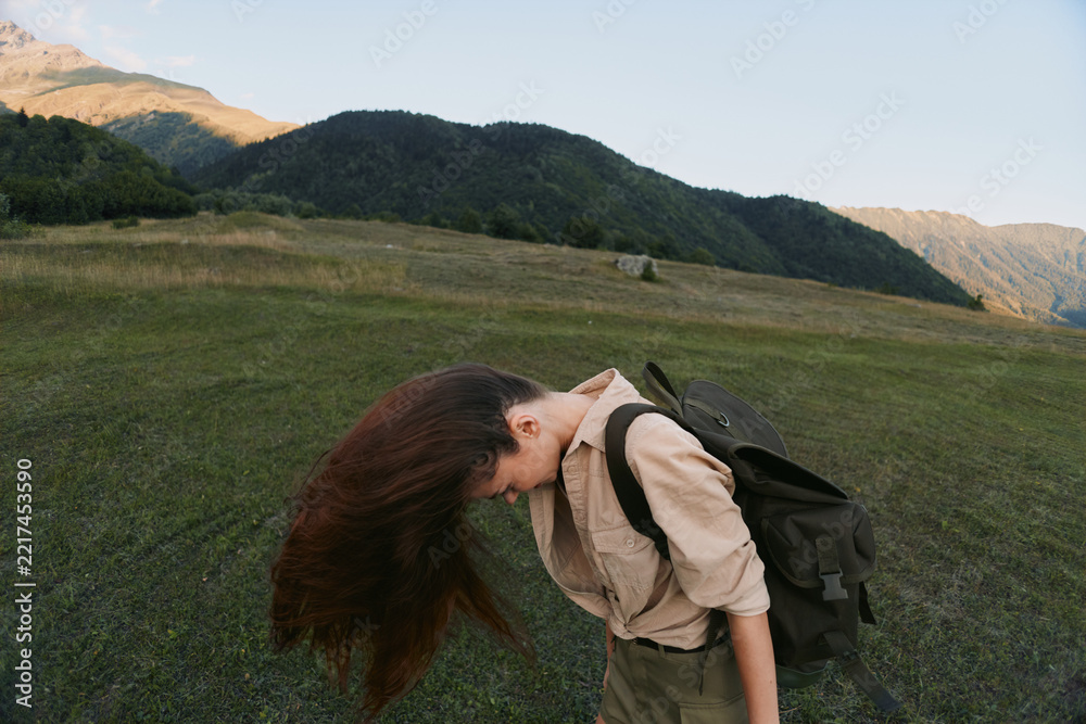 Naklejka premium Young woman outdoors bends forward in a grassy plain with a backpack, enjoying freedom and fresh air in a remote hillside landscape