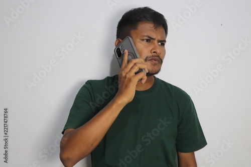 Young man talking on smartphone wearing green t shirt against white wall