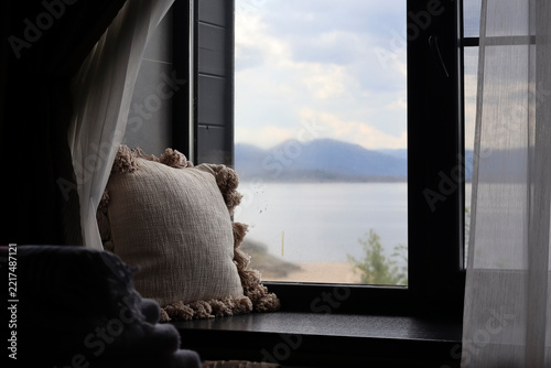Cozy interior with a soft decorative pillow by an open window overlooking a tranquil lake and distant mountains under a cloudy sky