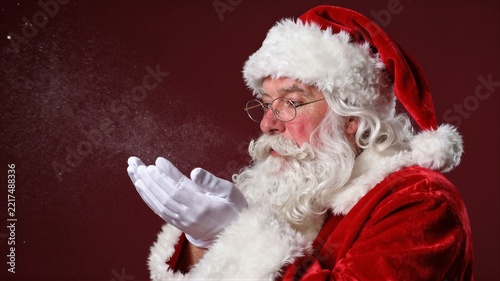 Elderly man in Santa costume blowing glitter from gloved hands against deep red background, with santa, santaclaus, santacostume, beard detail