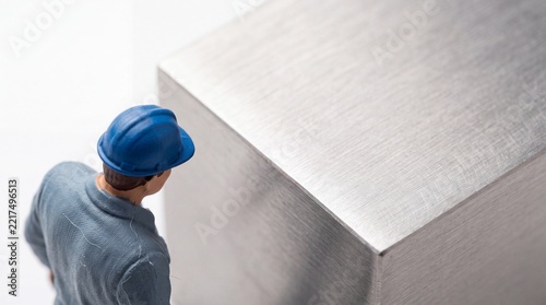 Miniature worker figure in a blue hard hat and grey uniform inspecting a large brushed stainless steel metal cube, macro concept for industrial quality control, manufacturing, and engineering.