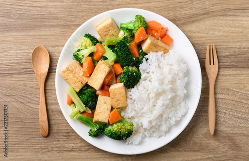 Stir fried tofu, broccoli, carrot and cooked rice in a white plate with fork and spoon on wooden background, Asian healthy vegetarian food, Table top view