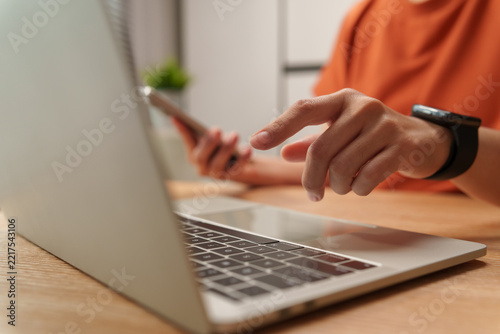 Woman using a smartphone beside a laptop while reaching toward the computer, capturing digital multitasking, connectivity and modern mobile productivity.