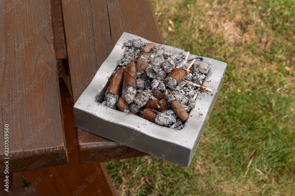 Fototapeta premium High angle view of a square stone ashtray filled with smoked cigarette butts and ash placed on an outdoor wooden table in daylight