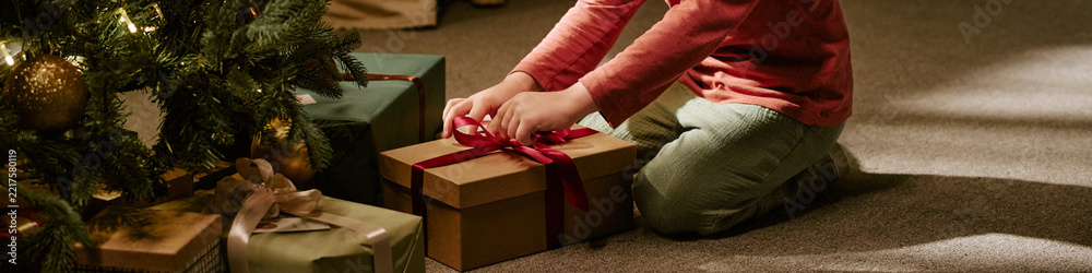 Fototapeta premium Header of Caucasian child opening gift box near Christmas tree at home. Image suitable for holiday marketing campaigns, seasonal retail promotions, family lifestyle advertising