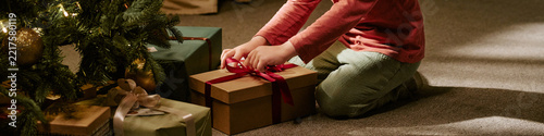 Header of Caucasian child opening gift box near Christmas tree at home. Image suitable for holiday marketing campaigns, seasonal retail promotions, family lifestyle advertising