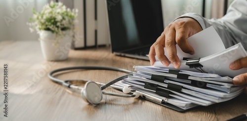 Stethoscope resting beside stacked documents while doctor manages paperwork, supporting healthcare records, medical administration, patient files and document workflow in clinic office.